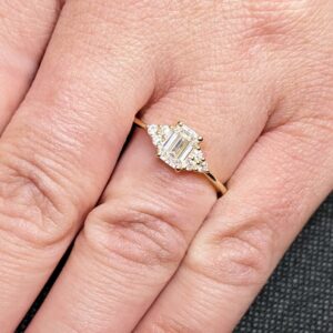 Delicate emerald-cut diamond engagement ring with side stones, set in 14K yellow gold, vintage-inspired minimalist design with elegant sparkle