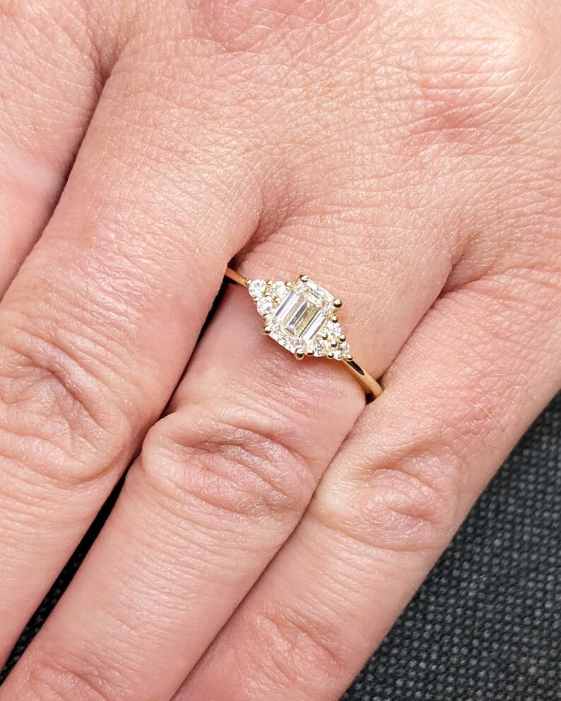 Delicate emerald-cut diamond engagement ring with side stones, set in 14K yellow gold, vintage-inspired minimalist design with elegant sparkle